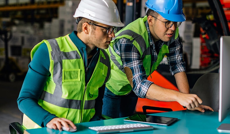 ROI calculator for manufacturing business efficiency Two manufacturing employees in safety gear reviewing data on a computer in a production facility