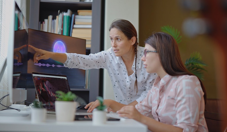 AI in B2B eCommerce: Team Collaboration on Data-Driven Strategy Two women collaborating in an office while analyzing AI-driven data visualizations on a screen, illustrating AI use in B2B eCommerce.