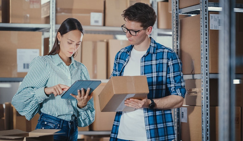 B2B Ecommerce Strategy in Action: Warehouse Operations Two warehouse employees managing B2B ecommerce orders using a tablet and shipping box.