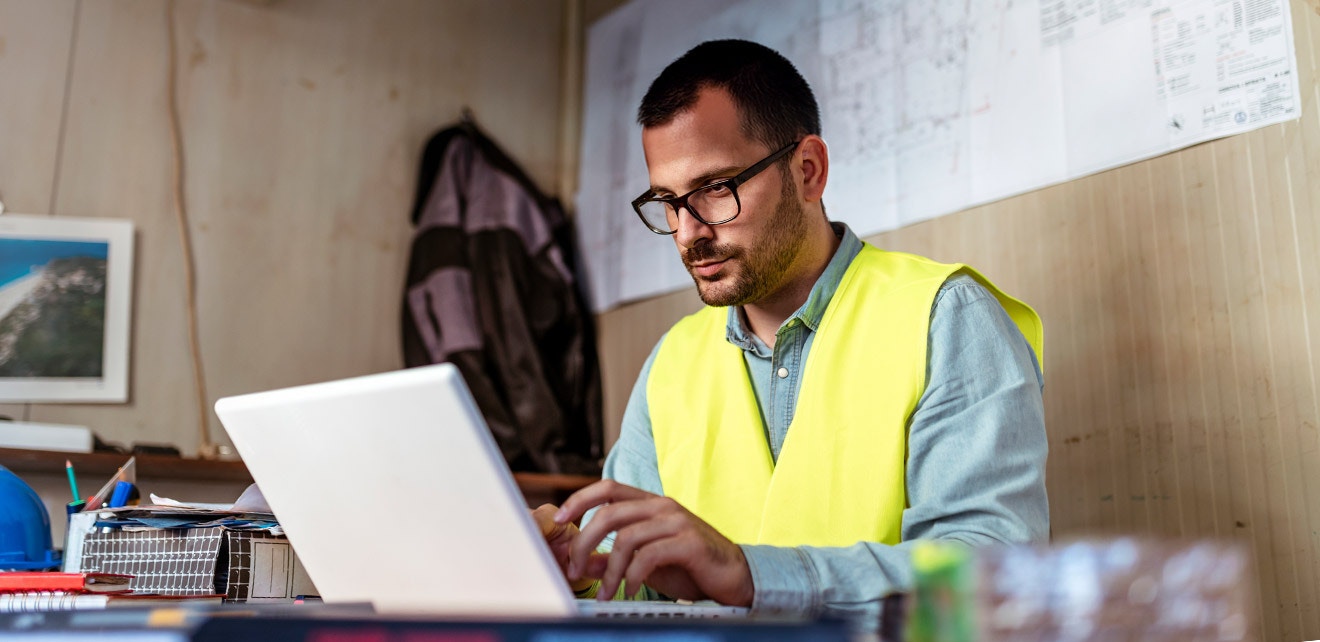 Construction Manager Using Trade Software Onsite A construction manager in a high-visibility vest working on a laptop in an office, using trade management software to oversee project tasks.