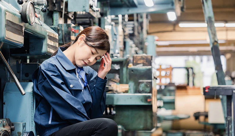 Eci blog Uk 10 signs erp implementation is failing banner An industrial worker in a blue uniform sits with her head in her hand, looking stressed and exhausted in a factory setting. She is surrounded by heavy machinery, emphasizing the high-pressure environment found in manufacturing or production facilities.