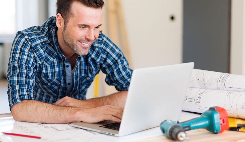 Homebuilding Contractor Using Project Management Software A smiling contractor reviewing homebuilding plans on a laptop, using trade contractor software at a job site.