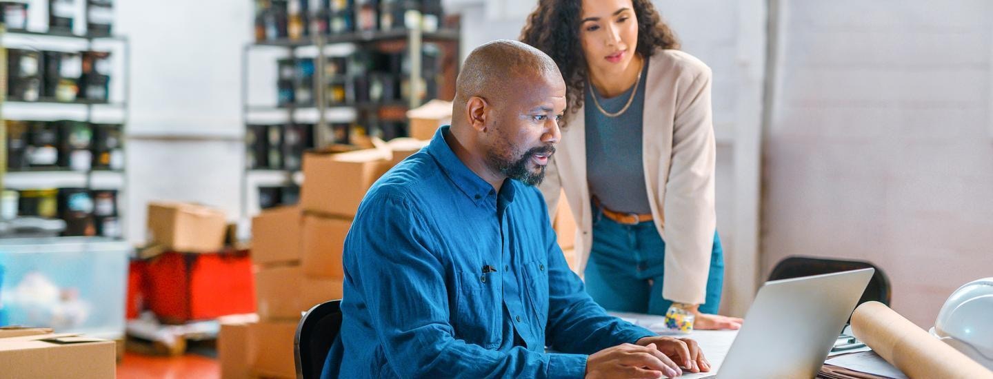 B2B Commerce Platform in Use at Office Tech Warehouse Two professionals using a laptop in a warehouse setting to manage office technology inventory via a B2B commerce platform.